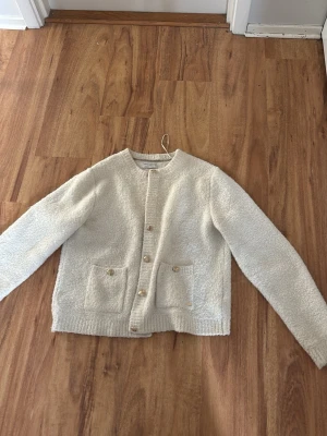 Vit teddykofta med knappar - Mysig vit teddykofta med rund halsringning och knäppning framtill. Koftan har två stora fickor framtill och ribbade muddar vid nederkant. Perfekt för lager-på-lager och en chill look.