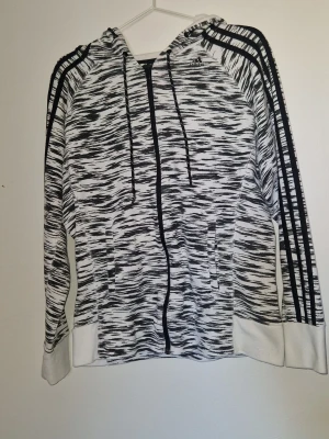Adidas kofta - Snygg hoodie från Adidas i svartvitt mönster med dragkedja framtill och klassiska svarta ränder längs ärmarna. Luva med snöre och två fickor framtill. Tillverkad i mjukt material som känns skönt mot huden. Perfekt för en sportig och avslappnad stil.