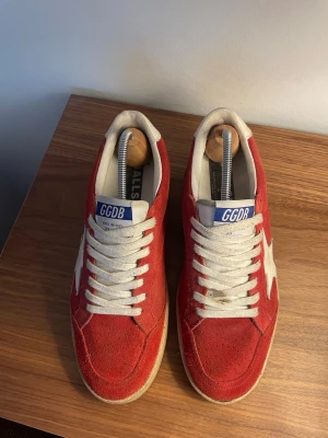 Röda GGDB sneakers med vit stjärna - Snygga röda sneakers från Golden Goose med vit stjärndetalj på sidan och vita skosnören. Ovandelen är i mocka och sulan är beige. Märkets logga syns på plösen i blått och vitt. Perfekta för dig som vill ha en unik och trendig look. Använda lite mer efter dessa bilder är tagna, äkthetsbevis finns.