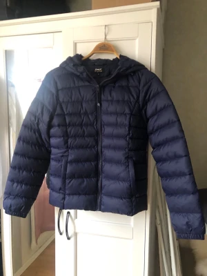 Mörkblå dunjacka från ONLY - Snygg mörkblå dunjacka från ONLY med huva och dragkedja framtill. Jackan har quiltad design och är perfekt för kalla vinterdagar. Lätt och smidig att bära, passar till många olika stilar.