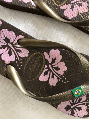 Bruna Havaianas sandaler med blommor - Säljer ett par bruna flip-flops från Havaianas med metallic-guldiga remmar och rosa hibiskusblommor på sulan. Klassisk brasiliansk flagga på remmen. Tillverkade i slitstark gummi, perfekta för sommaren eller stranden. Lite använda.