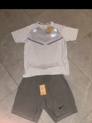 Nike träningsset t-shirt och shorts - Nike träningsset med en vit t-shirt med grått mönster och grå detaljer samt ett par grå shorts. T-shirten har rund hals och korta ärmar, båda plaggen har Nike-logga. Tillverkade i lätt och ventilerande funktionsmaterial, perfekt för träning.