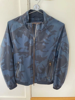 Blå camo vindjacka från Woolrich - Snygg vindjacka från Woolrich i blått camomönster. Jackan har dragkedja framtill, två fickor med dragkedja och hög krage. Perfekt för dig som gillar streetstyle och vill ha en lätt jacka med coolt mönster.