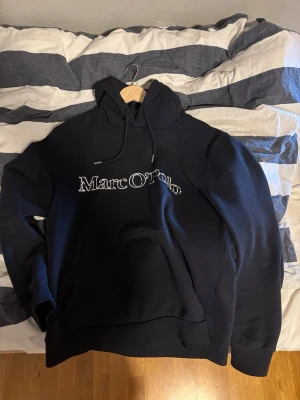 Svart hoodie från Marc O'Polo - Snygg mörkblå hoodie från Marc O'Polo med stor vit logga broderad på bröstet. Klassisk modell med huva och dragsnören samt en stor magficka framtill. Perfekt för en avslappnad och trendig stil. Nypris 1300.