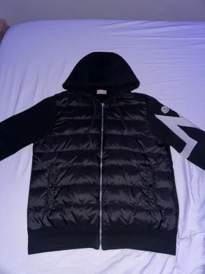 Svart pufferjacka med huva Moncler - Snygg svart pufferjacka från Moncler med huva och dragkedja framtill. Jackan har stickade ärmar med en vit blixtdetalj på vänster ärm och klassisk Moncler-logga. Kroppen är quiltad och vadderad för extra värme, perfekt för kalla dagar.