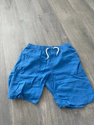 Blå shorts med snörning - Snygga blå shorts med elastisk midja och vit snörning framtill. Shortsen har sidofickor och två bakfickor, perfekta för sommardagar. Tillverkade i ett lätt och luftigt material som känns skönt mot huden.