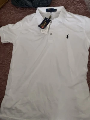 Vit pikétröja från Polo Ralph Lauren - Klassisk vit pikétröja från Polo Ralph Lauren med korta ärmar och knappslå framtill. Tröjan har den ikoniska broderade loggan på bröstet och är tillverkad i mjuk bomull. Perfekt till jeans eller shorts för en clean look.