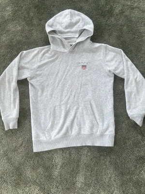 Ljusgrå hoodie från GANT - Snygg ljusgrå hoodie från GANT med klassisk huva och magficka. Diskret GANT-logga och sköld på bröstet i vitt och rött. Tillverkad i mjukt bomullsmaterial som är perfekt för chill dagar. Ribbade muddar vid ärmslut och nederkant för extra komfort.