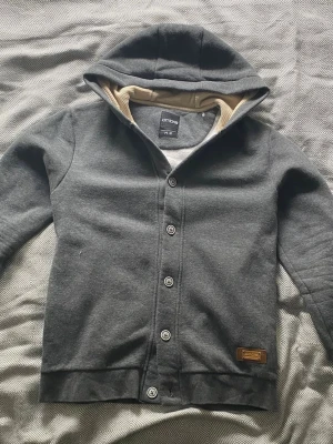 Mörkgrå hoodie med knappar från Ombre - Snygg mörkgrå hoodie från Ombre i storlek M med knappstängning framtill och beige innerfoder i huvan. Hoodien har en klassisk passform, ribbade muddar och en liten läderpatch vid nederkanten. Perfekt för en avslappnad och stilren look.