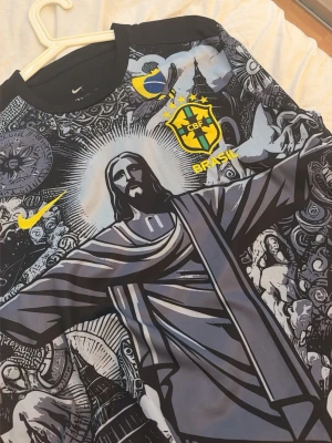 Nike Brasil CBF grafisk fotbollströja - Unik svart och grå fotbollströja från Nike med Brasilien CBF-emblem och grafiskt tryck av Kristusstatyn och brasilianska motiv. Tröjan har rund hals, gula detaljer och är tillverkad i ett lätt, tekniskt material som passar perfekt till träning eller match.