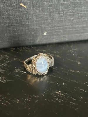 Silvrig ring med blå sten från mannasten - Unik silver ring med en oval, ljusblå sten i mitten. Ringen har en rå, organiskt formad design som ger ett coolt och handgjort intryck. Perfekt för dig som gillar statement-smycken med en konstnärlig touch. 3 cm i diameter 