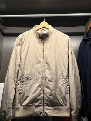 Beige bomberjacka från J.Lindeberg - Stilren beige vindjacka från J.Lindeberg med dragkedja framtill, två snedställda fickor och ribbade muddar vid ärmslut och nederkant. Jackan har en diskret logotyp på ärmen och är tillverkad i ett lätt material som passar perfekt för blåsiga dagar. Helt ny bara använd 1 gång. Det är storlek m men den passar S mer  