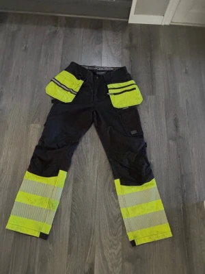Arbetsbyxa med reflex från Bluewear - Svarta arbetsbyxor med starka gula och reflexdetaljer från Bluewear. Byxorna har flera praktiska fickor framtill och förstärkta knän. Perfekta för dig som vill synas och behöver tåliga byxor till jobbet. Materialet är slitstarkt och designen är funktionell. Knappt använda.
