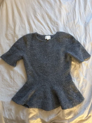 Grå stickad topp med volang - Mysig grå stickad topp med korta ärmar och volang nedtill. Tröjan har rund halsringning och är gjord i ett mjukt, fluffigt material. Tröjan är från HM är i bra skick. Kan skicka fler bilder om de frågas💛