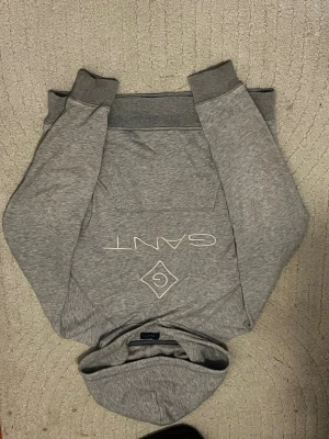Grå hoodie från Gant med logga - Snygg grå hoodie från Gant med vit logga och diamantmönster på bröstet. Klassisk känguruficka framtill och ribbade muddar vid ärmar och midja. Tillverkad i mjukt material och har en skön huva. Perfekt för en avslappnad och trendig look.
