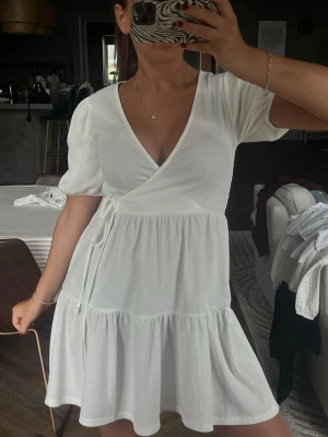 Vit omlottklänning med knyt - Supersöt vit omlottklänning med korta puffärmar och knyt i midjan. Klänningen har v-ringning och volangkant nedtill för en extra feminin touch. Perfekt för varma dagar och enkel att styla med smycken eller sneakers.