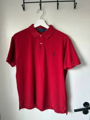 Röd pikétröja från Polo Ralph Lauren - Klassisk röd pikétröja från Polo Ralph Lauren med blå broderad logga på bröstet. Tröjan har korta ärmar, krage och två knappar framtill. Tillverkad i bomull för en bekväm och luftig känsla. Priset ej hugget i sten! Kontakta vid frågor:)