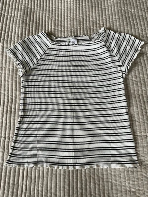 Randig ribbad t-shirt i vitt och svart  - Säljer en kortärmad t-shirt med svartvita ränder och ribbad struktur. T-shirten har en lätt croppad passform och små volangdetaljer vid ärmslut och hals. Perfekt till jeans eller kjol.