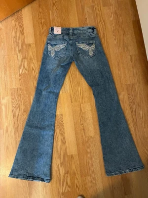 Blå bootcut jeans med paljettfickor - Säljer ett par blå bootcut jeans med snygga broderade bakfickor dekorerade med paljetter i silver och beige. Jeansen har låg midja och klassisk femficksdesign. Perfekta för dig som gillar detaljer och retrostil.