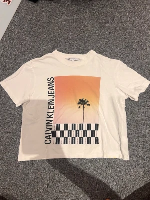 Vit t-shirt Calvin Klein Jeans S - Vit t-shirt från Calvin Klein Jeans med tryck av palm, grafiskt mönster och text på framsidan. T-shirten har korta ärmar och rund hals. Materialet är mjuk bomull och passformen är normal. Perfekt för en chill och stilren look.