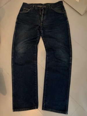 Mörkblå raka jeans från Wrangler - Säljer ett par mörkblå jeans från Wrangler med rak passform och klassisk femficksdesign. Jeansen har en snygg tvätt och är tillverkade i slitstark denim. W34 L32 skriv privat för mått. De är väl använda men i fint skick, finns en liten slitning på baksidan som visat i bilden men inget att oroa sig för. Midja: 44cm längd: 112cm