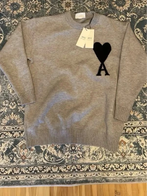 Grå stickad tröja från Ami Paris - Snygg grå stickad tröja från Ami Paris med svart hjärta och bokstaven A broderat på bröstet. Tröjan har rund halsringning, ribbade muddar och är tillverkad i mjukt material som passar perfekt till höst och vinter.