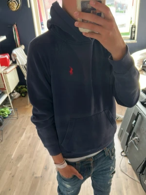 Mörkblå hoodie från Polo Ralph Lauren - Säljer en mörkblå hoodie från Polo Ralph Lauren med klassisk röd logga broderad på bröstet. Hoodien har huva, ribbade muddar och en stor magficka. Perfekt för dig som gillar stilren och sportig look.