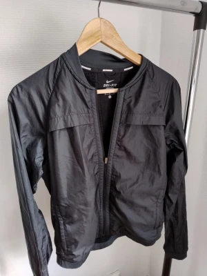 Svart löparjacka från Nike Dri-Fit - Svart löparjacka från Nike med Dri-Fit teknologi som håller dig torr under träningen. Jackan har långa ärmar, dragkedja framtill och meshpaneler för extra ventilation. Perfekt för löpning och andra sportaktiviteter.