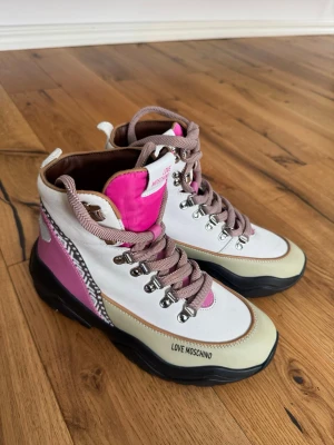 Love Moschino färgglada kängor - Säljer ett par trendiga kängor från Love Moschino i vitt, beige och rosa med svarta detaljer. Skorna har grov sula, metallhål för snörning och mönstrade snören. Materialet är en mix av textil och syntet. Perfekta för dig som vill sticka ut med din stil.