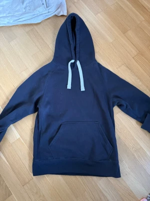 Russemerch hoodie - Mörkblå hoodie med stor känguruficka framtill och ljusblåa dragsnören i huvan. Tröjan har raglanärmar och broderad textdetalj på ena ärmen. Tillverkad i mjukt bomullstyg, perfekt för en avslappnad stil.