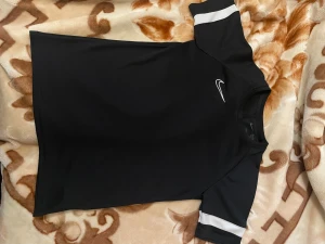 Svart Nike sporttröja med vita detaljer - Snygg svart sporttröja från Nike med vita ränder på ärmarna och klassisk Nike-logga på bröstet. Tillverkad i ett lätt och snabbtorkande material som passar perfekt till träning eller sportaktiviteter. Kortärmad och med rund hals.