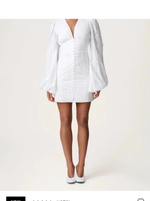 Vit kort klänning från adoore - Stilren vit kort klänning med v-ringning och långa puffärmar. Klänningen har knappar framtill och är sydd i ett lätt material med diskret struktur. Perfekt för dig som vill ha en elegant och trendig look.