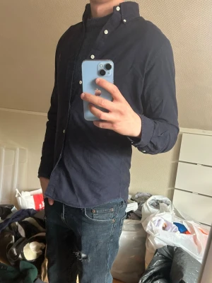 Mörkblå skjorta från Ralph Lauren - Säljer en mörkblå skjorta från Ralph Lauren med klassisk button-down krage och vita knappar. Skjortan har lång ärm och den ikoniska röda logotypen broderad på bröstet. Tillverkad i bomull och har en normal passform.