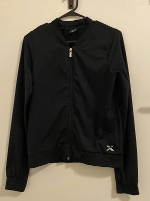 Svart träningsjacka från Xtra - Snygg svart träningsjacka från Xtra med dragkedja framtill och diskret logga nere vid höften. Jackan har långa ärmar, ribbade muddar och är tillverkad i ett lätt och smidigt material som passar perfekt till träning eller uppvärmning.
