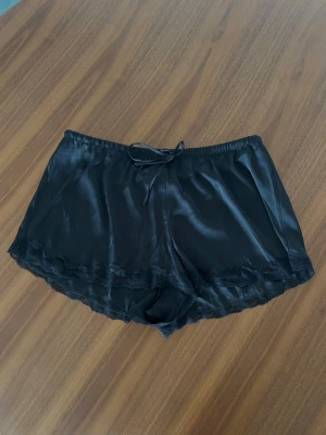 Svarta satinskjorts med spetskant - Helt nya med prislapp🤍 säljer även matchande topp om man vill ha ett set🤍Snygga svarta loungewear-shorts i glansigt satinmaterial med dekorativ spetskant längst ner. Shortsen har resår i midjan och en liten rosett framtill för extra detalj. Perfekta för en chill dag hemma eller mysiga kvällar.