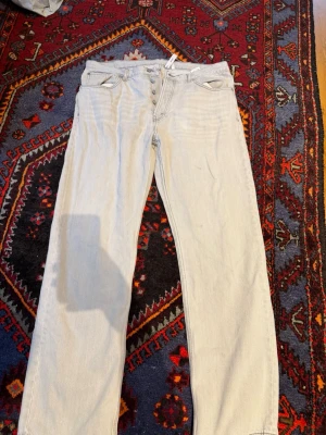 Ljusblå raka jeans med knappar - Säljer ett par ljusblå jeans med raka ben och knappgylf. Jeansen har klassisk femficksdesign och är tillverkade i denim. Perfekta för en avslappnad och trendig look.