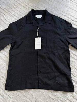Svart linneskjorta från Zara - Stilren svart skjorta från Zara i luftig linnemix. Skjortan har klassisk krage, knäppning framtill och korta ärmar. Perfekt för en avslappnad och trendig look. 100% Linne