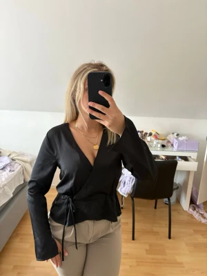 Svart  satin omlottblus med knytband - Stilren svart omlottblus med långa ärmar och knytband i midjan. Blusen har djup v-ringning och en lätt glansig finish som ger en elegant touch. Perfekt att matcha med både jeans och kjol för en trendig look. Prislapp kvar. Nypris 400kr