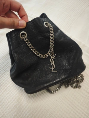 Svart bucket bag  - Säljer en svart bucket bag i mjukt läder med grov silverfärgad. Väskan har dragskoöppning och cool metallkedja som axelrem. Perfekt för dig som gillar en edgy och lyxig look.