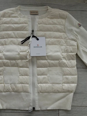 Vit quiltad cardigan från Moncler - Stilren vit cardiganjacka från Moncler med quiltat mönster framtill, två fickor och dragkedja. Långa ärmar i mjukt stickat material och rund halsringning. Moncler-logga på ärmen ger en exklusiv touch.