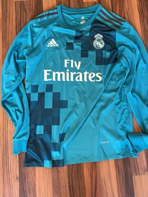 Real Madrid 2017 borta tröja - Snygg långärmad Real Madrid matchtröja från Adidas i turkosblått med pixelmönster i mörkblått. Tröjan har klubbmärke och Adidas-logga på bröstet samt Fly Emirates-tryck. Tillverkad i lätt och ventilerande material med Climalite-teknologi. 