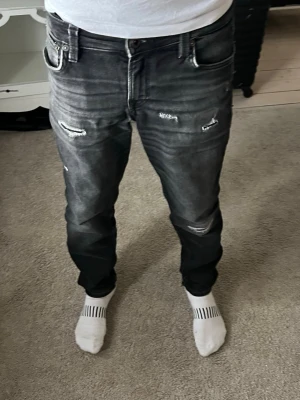  Jack & Jones Slim Glenn jeans - Säljer ett par svarta jeans från Jack & Jones med slitningar och distressed detaljer på framsidan. Jeansen har klassisk femficksdesign och raka ben. Materialet är denim med en cool tvätt som ger en trendig look.