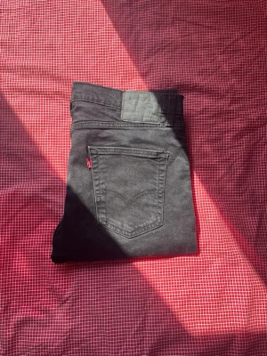 Svarta Levi's jeans med rak passform - Säljer ett par svarta jeans från Levi's med klassisk rak passform och femficksmodell. Jeansen har den ikoniska röda Levi's-lappen på bakfickan och är tillverkade i ett mjukt denimtyg. Perfekta för en clean och stilren look.