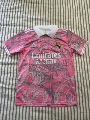 Rosa Real Madrid tröja med drakmotiv - Snygg rosa Real Madrid matchtröja från Adidas med unikt grått drakmotiv över hela plagget. Tröjan har vit krage, vita detaljer på ärmarna och klubbmärke samt Adidas-logga på bröstet. Tillverkad i lätt och ventilerande funktionsmaterial.
