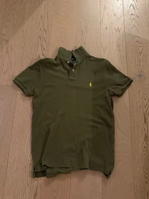 Olivgrön pikétröja från Ralph Lauren - Klassisk olivgrön pikétröja från Ralph Lauren med korta ärmar och krage. Tröjan har två knappar vid halsen och den ikoniska gula logotypen broderad på bröstet. Tillverkad i mjuk bomull för en bekväm känsla.