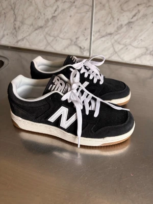 Svarta New Balance sneakers - Snygga svarta New Balance sneakers med vit sula och vita detaljer. Skorna har klassisk låg profil, vit logga på sidan och vita skosnören. Ovandelen är i mocka och mesh för en sportig och stilren look. Perfekta för dig som gillar streetwear och retrostil.