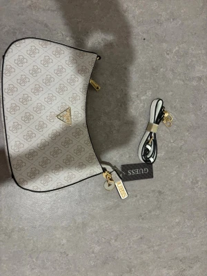Vit axelväska från Guess - Snygg vit handväska från Guess med beige mönster och guldfärgade detaljer. Väskan har en avlång form, svart kant och medföljande axelrem. Materialet är konstläder och väskan har Guess-logga i guld på framsidan.