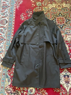 Svart trenchcoat MarcOPolo - Stilren svart trenchcoat med hög krage och knäppning framtill. Klassisk stormflik över bröstet och justerbara remmar vid ärmsluten. Två snedställda fickor och rak passform. Perfekt för en trendig och tidlös look.
