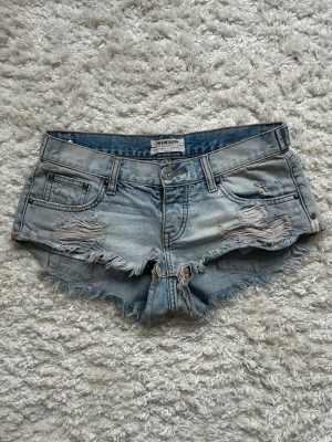Ljusblå slitna jeansshorts One Teaspoon - Säljer ett par ljusblå jeansshorts från One Teaspoon med slitna detaljer, fransig kant och klassisk femficksmodell. Shortsen har låg midja och är perfekta för sommaren💕 Rå och trendig look med knapp framtill.