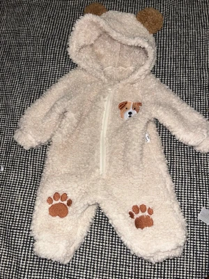 Beige teddyoverall med hundmotiv - Mysig beige teddyoverall med huva och söta björnöron. Dragkedja framtill, broderad hund på bröstet och tassavtryck på benen. Perfekt för att hålla sig varm och gullig under kalla dagar. Tillverkad i fluffigt, mjukt material.